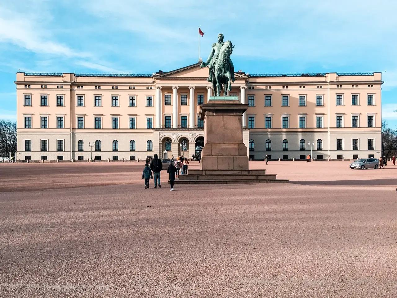 Front exterior of the Oslo Royal Palace, with the Norwegian flag flying above the building and a statue of a man on a horse in front of the palace.