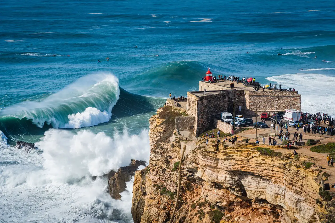 Onlookers standing at the Nazare Red lighthouse overlooking huge waves and surfers.