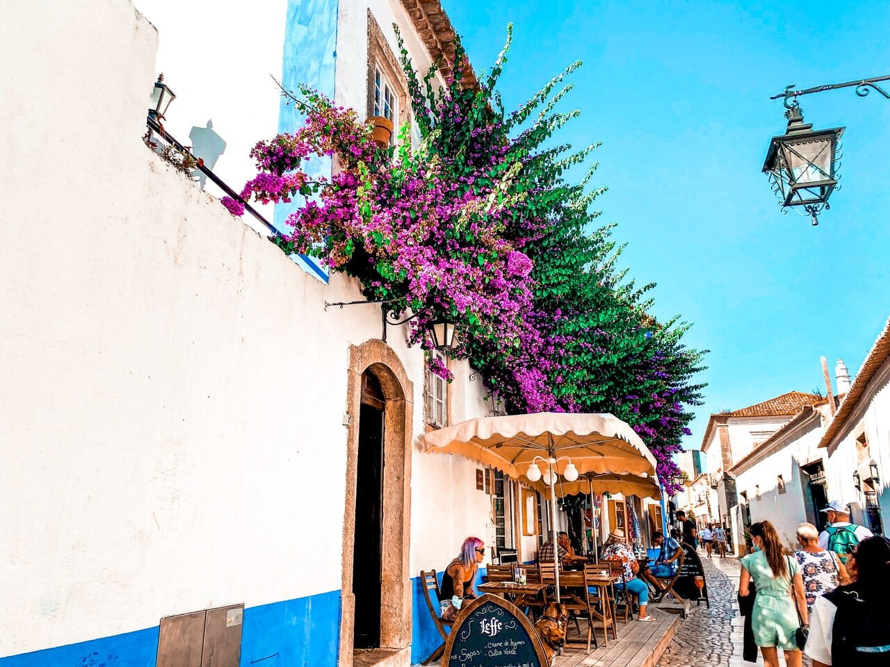 Cobbled street in the medieval town of Óbidos, Portugal. People are sitting outside a restaurant in the sun whilst others stroll by.