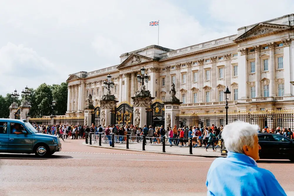 The front gates at Buckingham Palace, swarmed with tourists, with an iconic London black cab driving past. Buckingham Palace is one of the best palaces in London to visit.