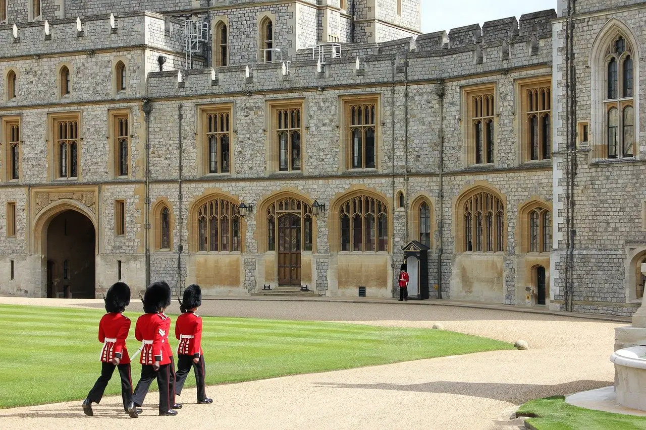 Windsor Castle, a royal castle in London England. Four soldiers in red coats and black bearskin hats march in the grounds as an additional soldier stands guard. It is an official residence of the British Royal Family.