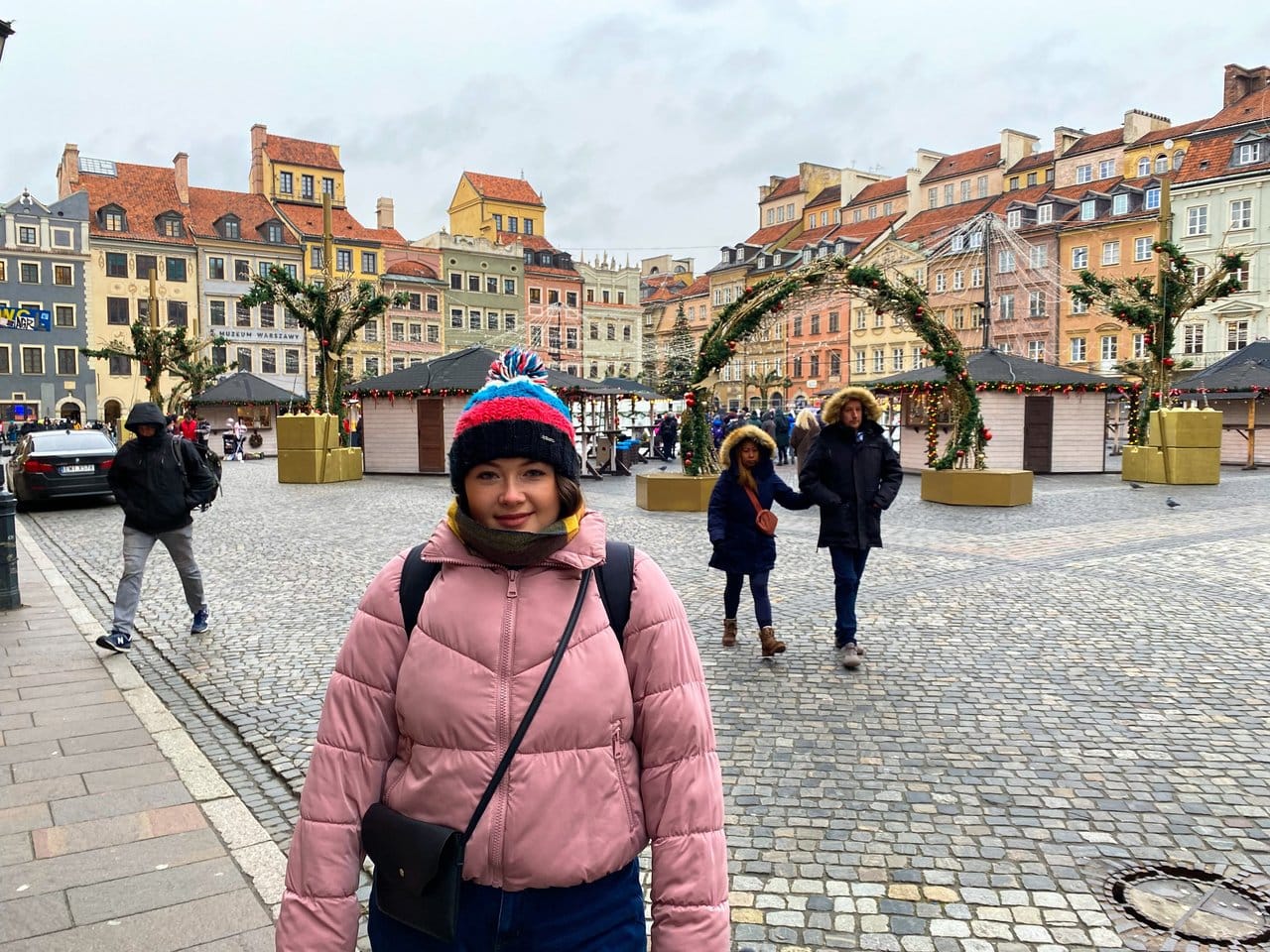 Ella wearing a pink coat and hat whilst travelling in Poland with the Warsaw Christmas Markets in the background. These are some of the cheapest Christmas Markets in Europe.