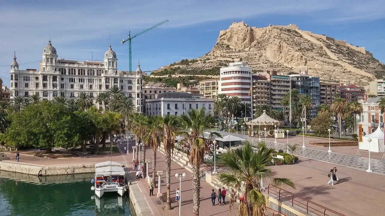 The port of Alicante, one of the best cities in Spain. A small ferry is in the water, which is next to a pedestrian walkway. In the background, you can see a rocky hill with a castle on top and a regal building.