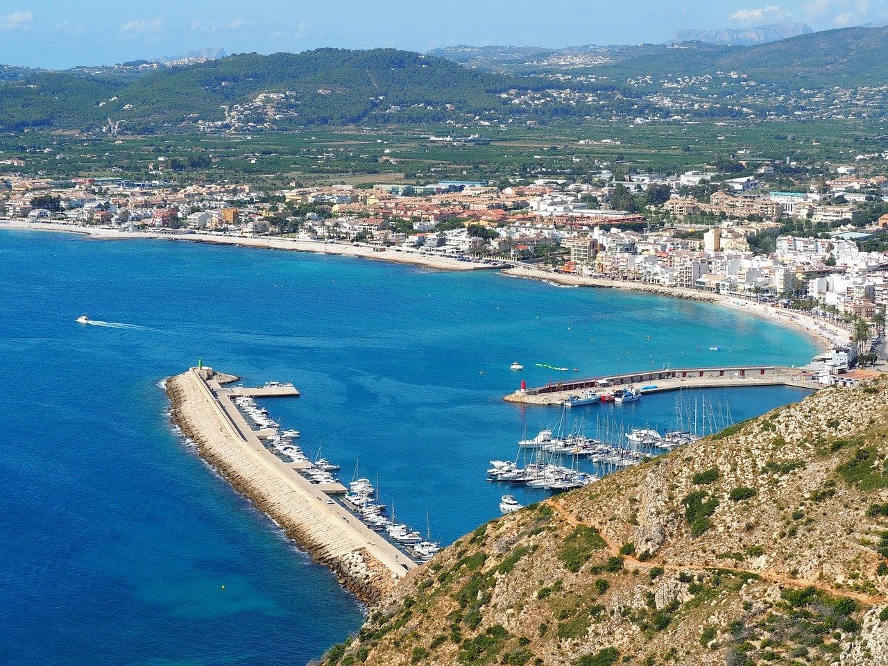 The port of Jávea/Xàbia in the Costa Blanca. On the right side you can see Javea harbour, with Javea's beaches in the background. Buildings are dotted along the coastline.