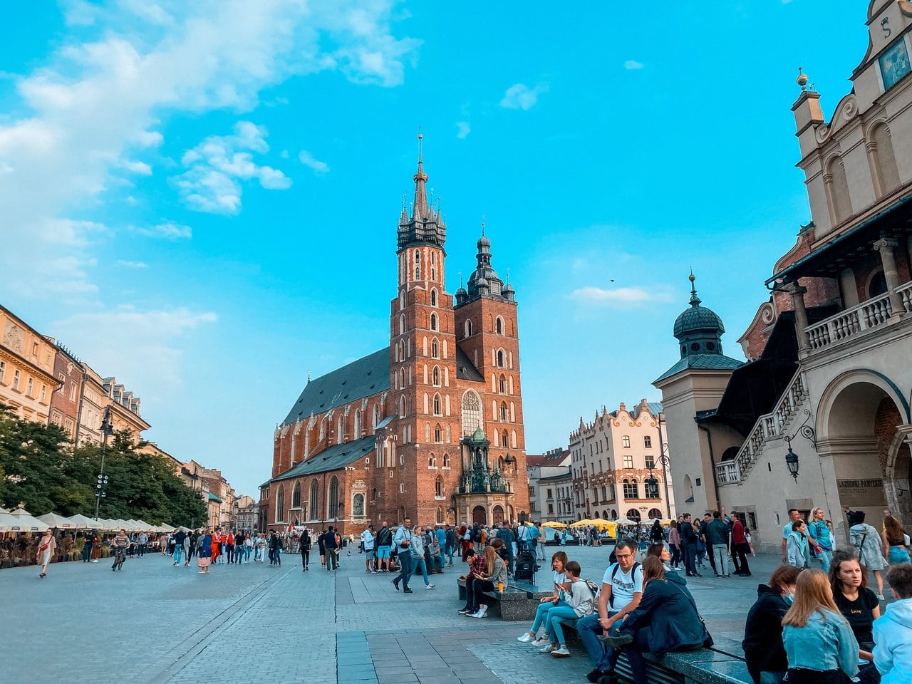 Crowds of people enjoy a sunny day during a weekend in Krakow, with the prominent twin spires of the Gothic-style St. Mary's Basilica towering in the background against a clear blue sky with scattered clouds.