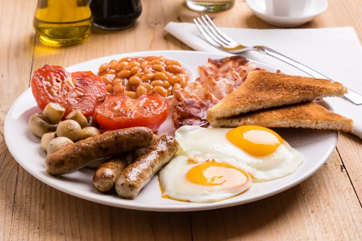 a Full English breakfast in Clapham, consisting of two fried eggs, three sausages, grilled tomatoes, toast, bacon, beans and mushrooms on a white plate.