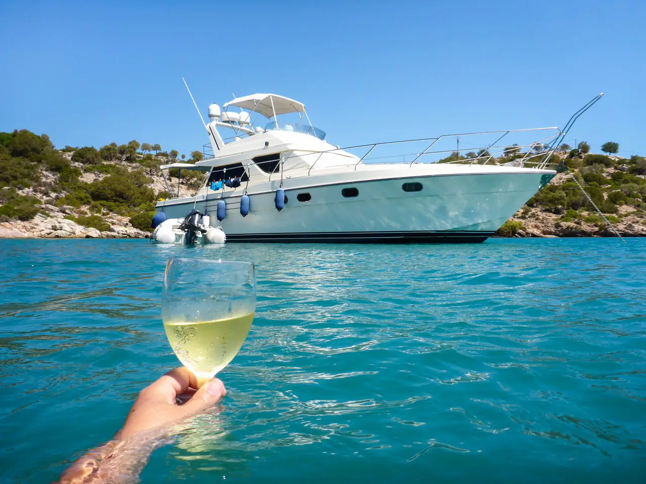 Person in blue water holding a glass of white wine in front of a small yacht.