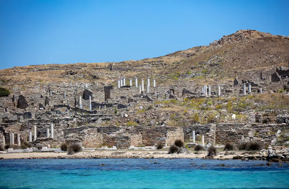 Delos Cyclades island, Greece. General view approaching by boat. Holy sanctuary archaeological site, UNESCO Heritage monument. Stone walls and marble pillars ruins, on rocky hill. Blue sky and sea