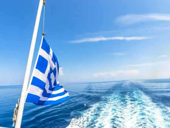 Greek flag hanging off the back of a boat fluttering in the wind. Sea and blue skies in the background.
