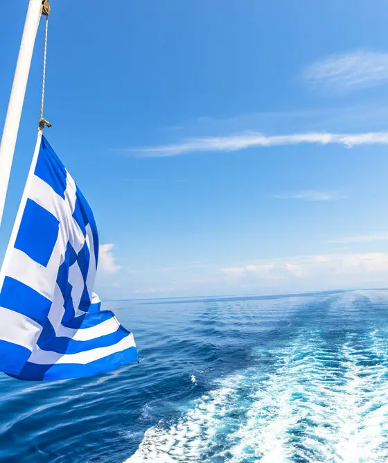 Greek flag hanging off the back of a boat fluttering in the wind. Sea and blue skies in the background.