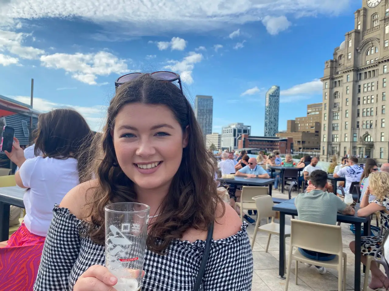 Ella, holding up a pint of beer at a Liverpool rooftop bar. City skyscrapers are in the background. Liverpool is one of the most affordable cities in the UK!