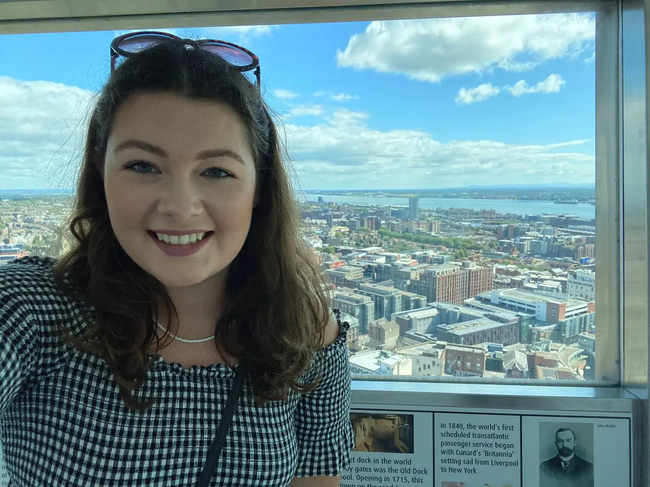 A selfie of Ella wearing a black and white gingham dress at the top of the St John's Beacon in Liverpool. The city skyline is in the background.