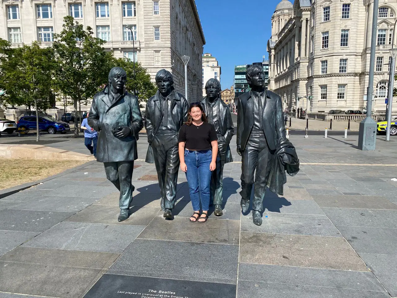 Ella, wearing blue jeans and a black top, posing with the Beatles sculpture in Liverpool. Liverpool is worth visiting for Beatles fans!