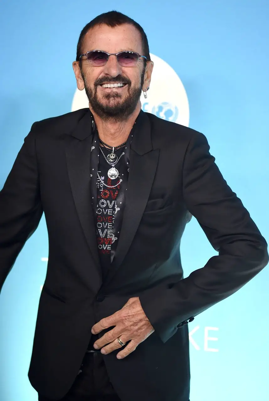 Ringo Starr in the present day standing against a blue backdrop wearing a black blazer and sunglasses. Ringo is the oldest Beatle.
