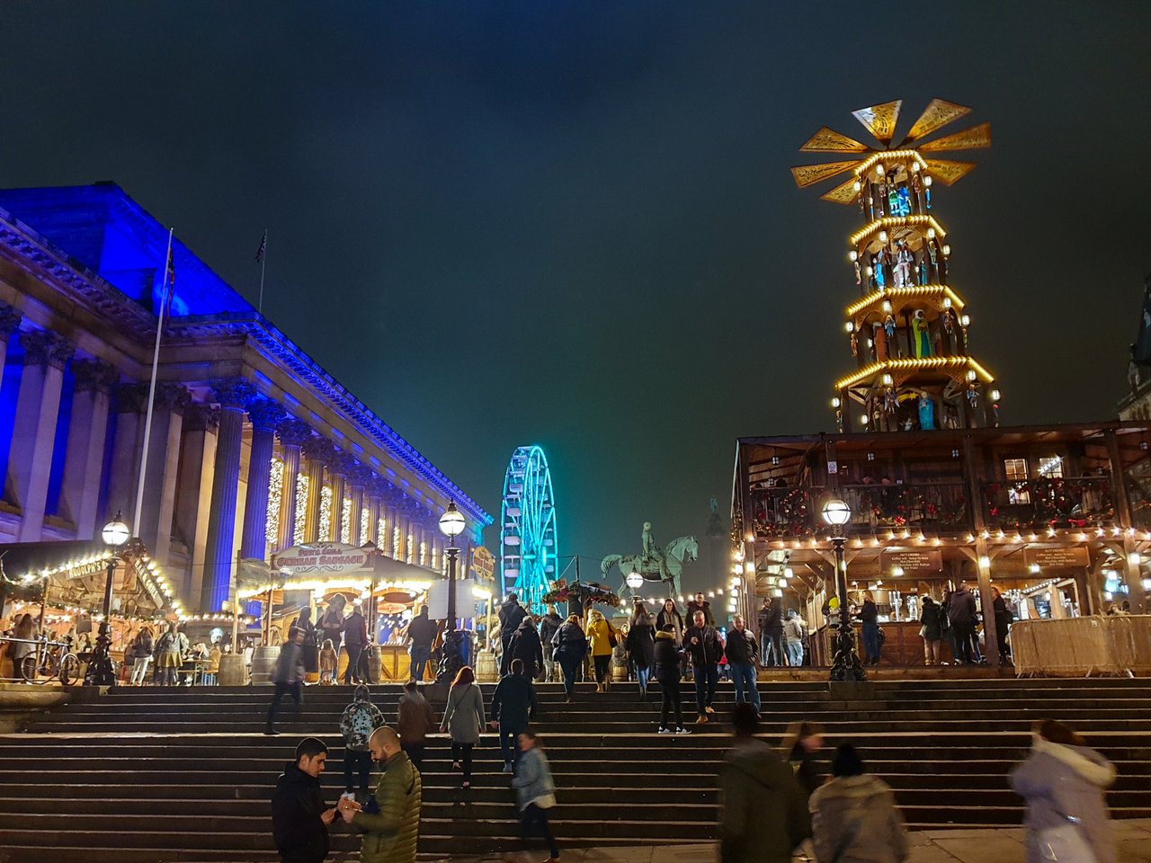 View of Liverpool Christmas market at night, with wooden huts, the illuminated St George's Hall, and a ferris wheel in the background