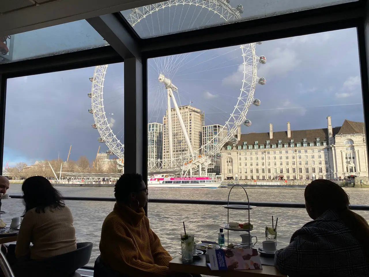 River Thames Afternoon Tea Boat Cruise in London. Afternoon tea trays are stacked on tables and you can see the London Eye from the boat's window