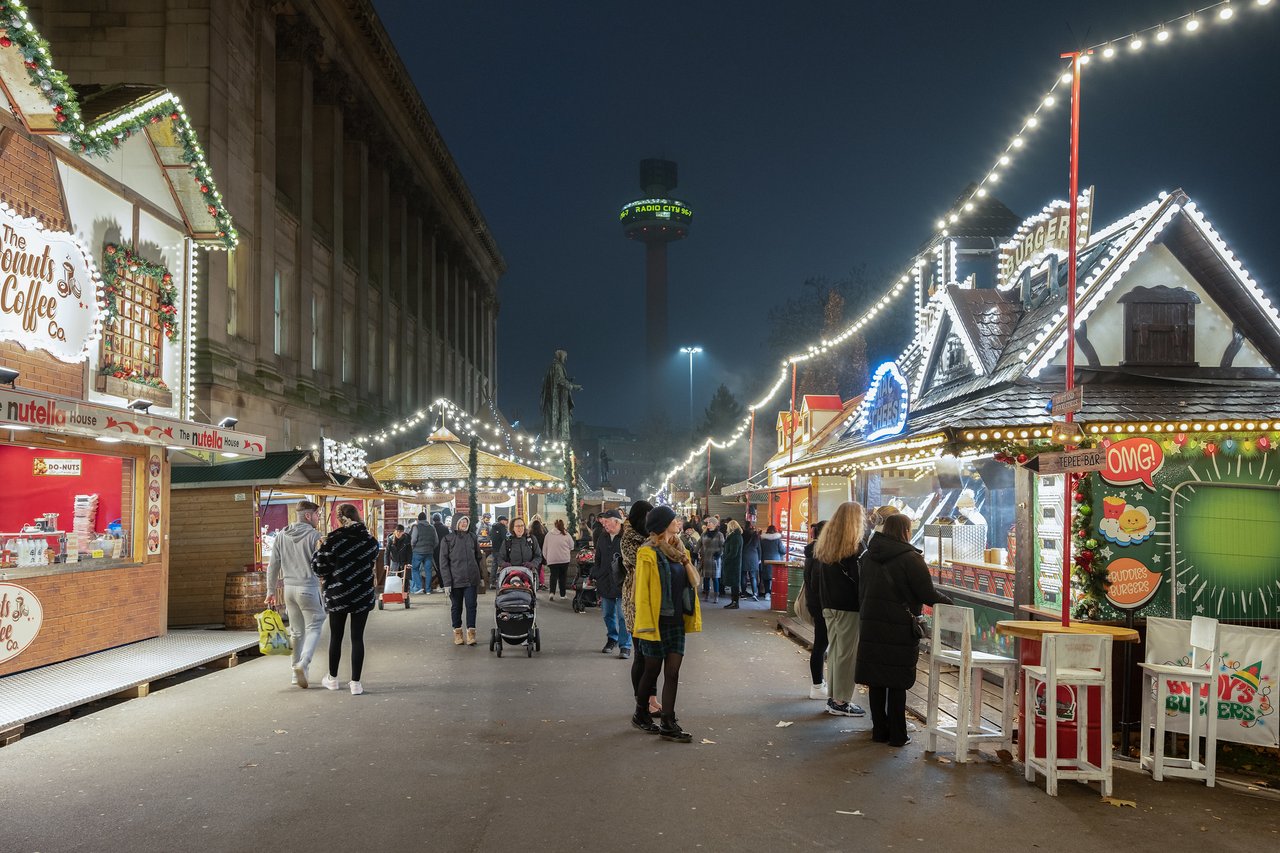 People browsing Christmas market stalls in Liverpool at night with the St John's Beacon in the background