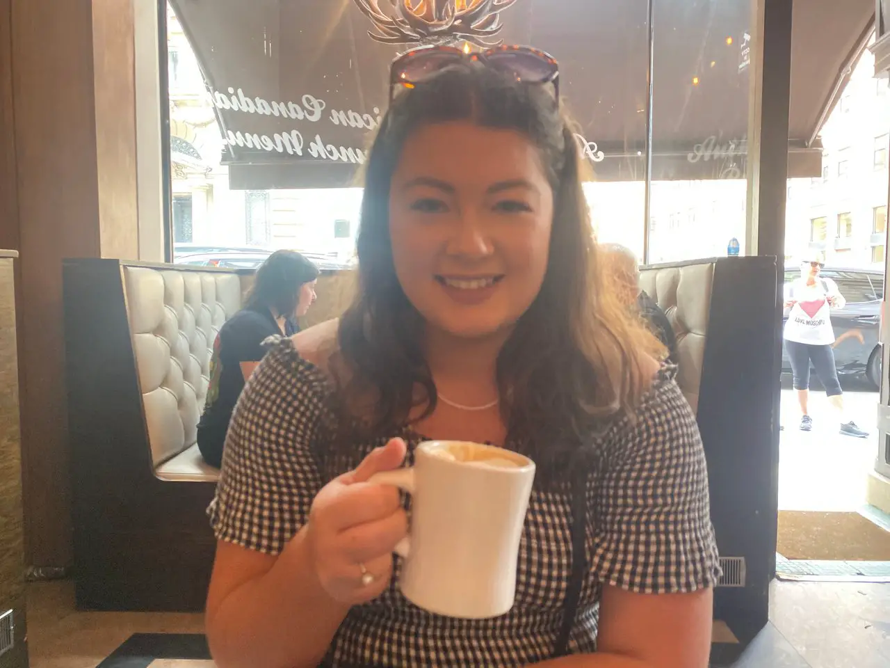 Ella wearing a black and white dress and sunglasses smiling with a cup of coffee whilst having brunch at Moose Coffee in Liverpool.