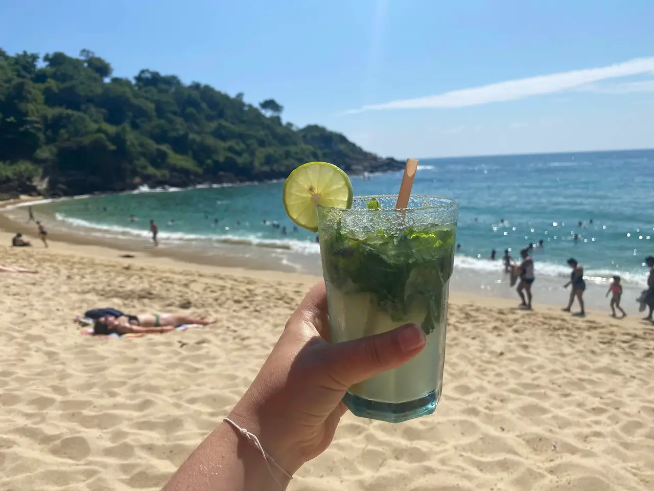 Person holding out a mojito with a lime wedge on a sandy beach in Mexico with the ocean in the background.