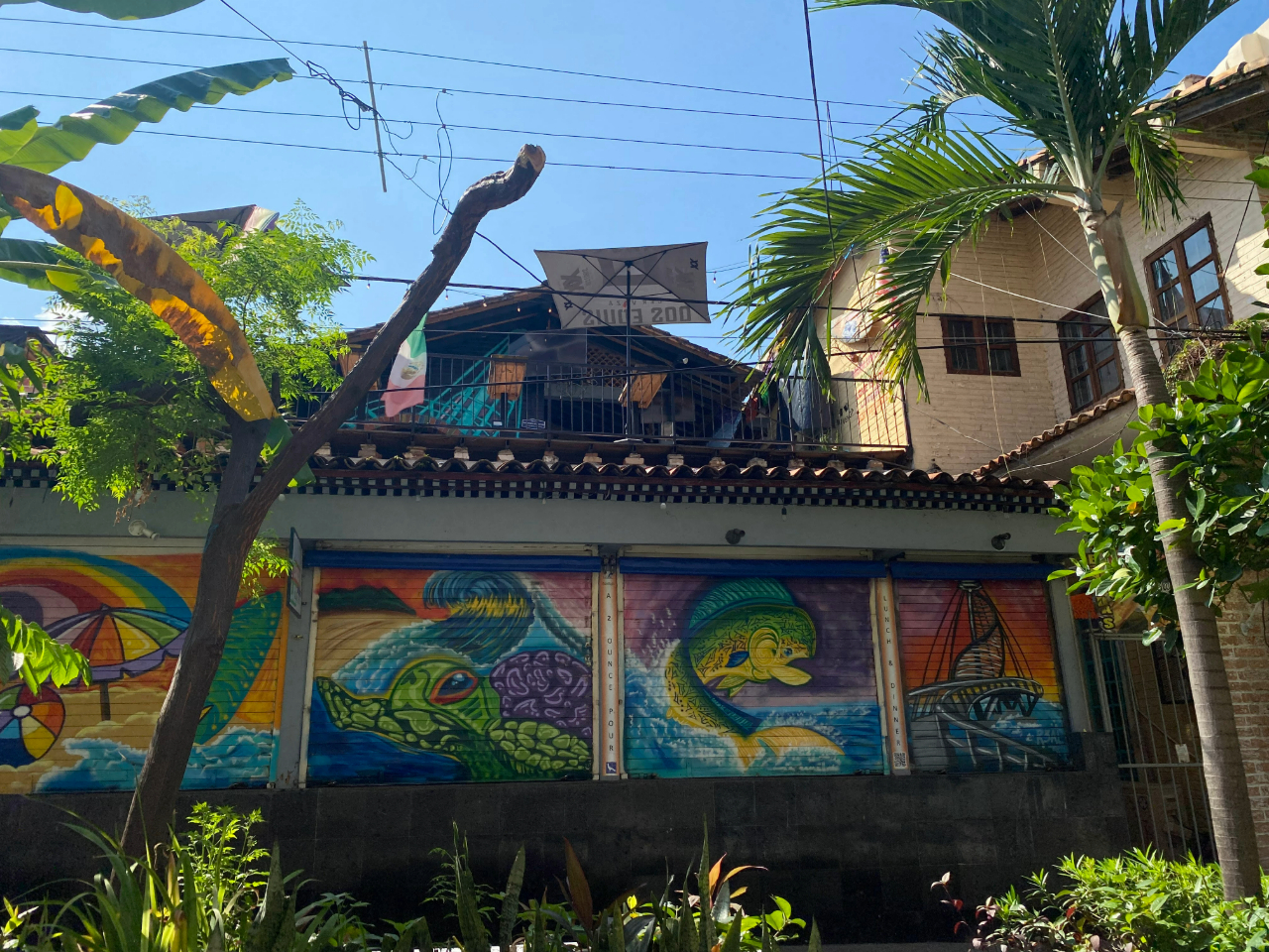  A colourful mural on a building in Jalisco, Mexico, featuring vibrant ocean-themed artwork, including a sea turtle, a large fish, and a beach scene with an umbrella. The upper level of the building has a rustic rooftop with a Mexican flag, satellite dishes, and exposed wiring, surrounded by tropical greenery.